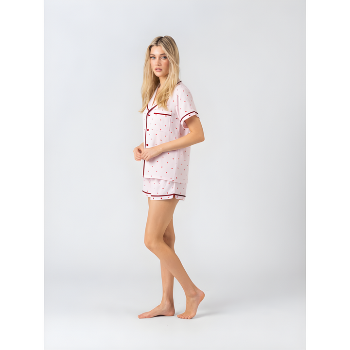 Side view of a woman wearing light pink short-sleeve pyjamas with red heart print and red piping trim.